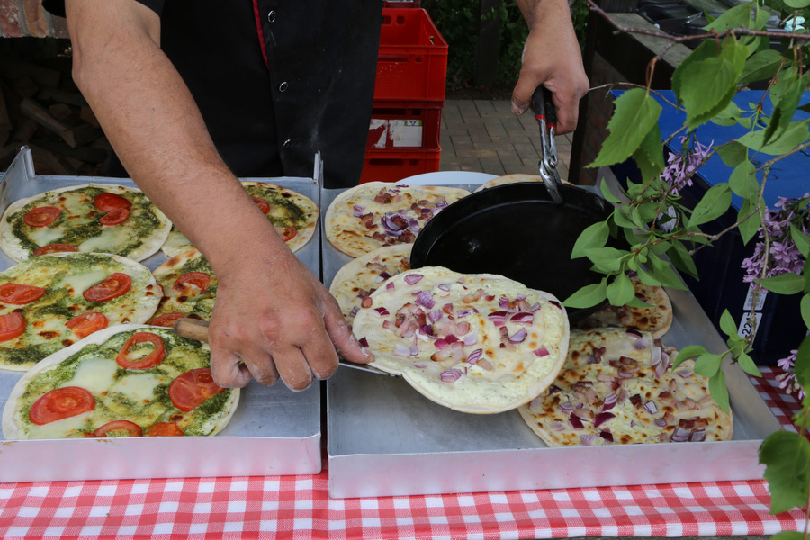 KI generiert: Eine Person bereitet Flammkuchen mit Zwiebeln und Tomaten auf einem Tisch mit rot-weiß kariertem Tuch zu.