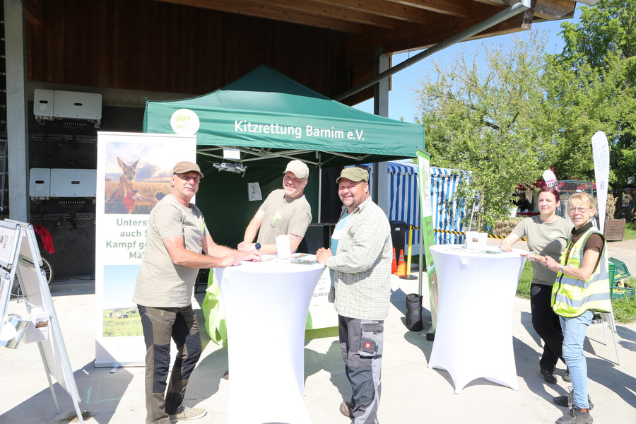 KI generiert: Eine Gruppe von Menschen steht an Stehtischen vor einem Stand mit der Aufschrift "Kitzrettung Barnim e.V.".