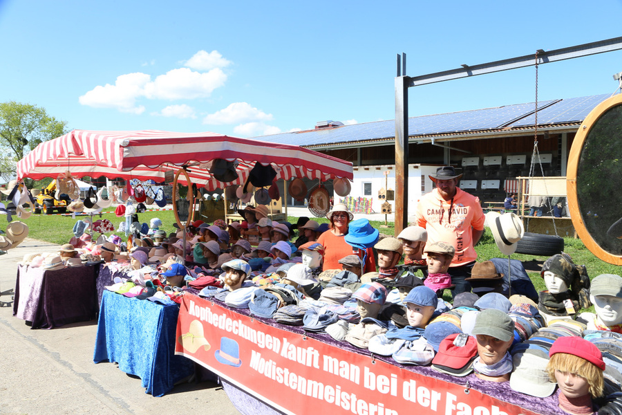 KI generiert: Ein Stand mit vielen Hüten unter einem rot-weißen Sonnenschirm. Text: "Kopfbedeckungen kauft man bei der Fach... Modistenmeisterin".