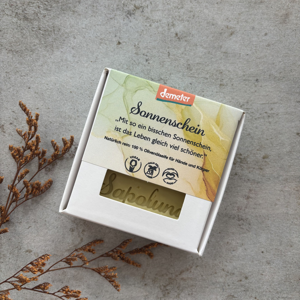 KI generiert: Verpackte Olivenölseife für Hände und Körper mit dem Namen "Sonnenschein". Text: „Mit so ein bisschen Sonnenschein...".