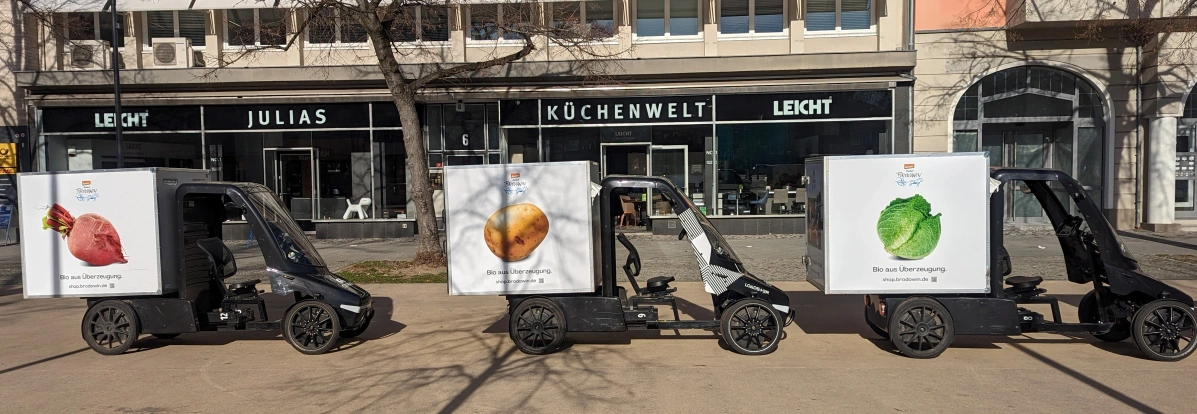 KI generiert: Vier Lieferwagen mit Gemüsebildern parken vor einem Küchenstudio. Texte: "Bio aus Überzeugung".