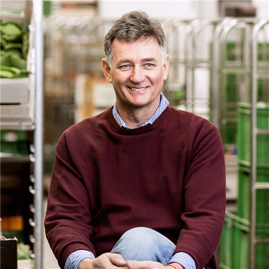 KI generiert: Zwei Personen stehen in einem Gewächshaus voller Pflanzen und halten eine Kiste mit frischem Gemüse. Der Fokus liegt auf der Präsentation der Ernte.