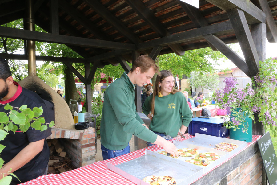 KI generiert: Zwei Personen servieren Pizza an einem Stand mit Backofen, im Freien, umgeben von Bäumen und Blumen.
