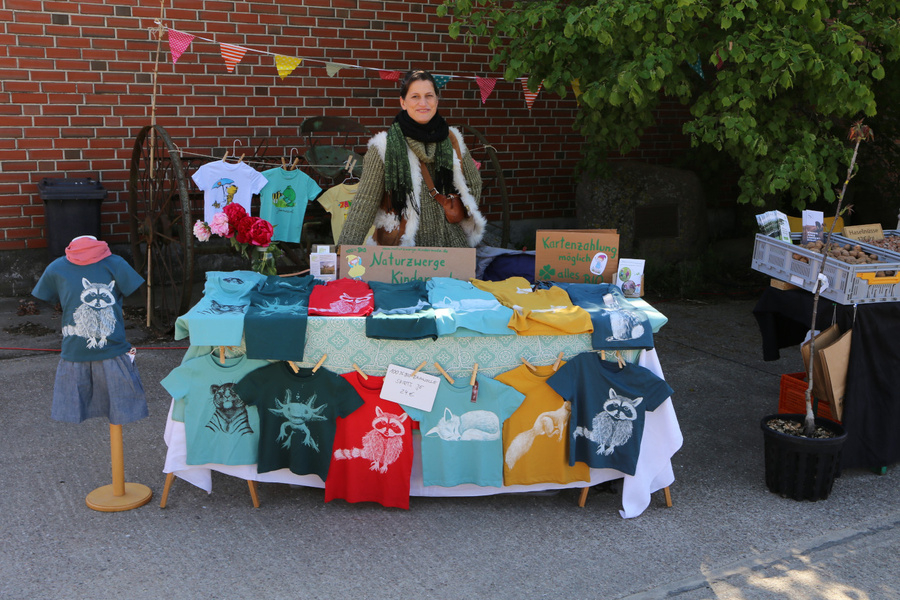 KI generiert: Frau an einem Marktstand mit bunten Kinder-T-Shirts, Schilder: "Naturzwerge Kleidung", "Kartenzahlung möglich".