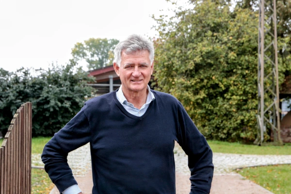 KI generiert: Ein Mann steht lächelnd im Freien, im Hintergrund sind Bäume und ein Gebäude zu sehen.