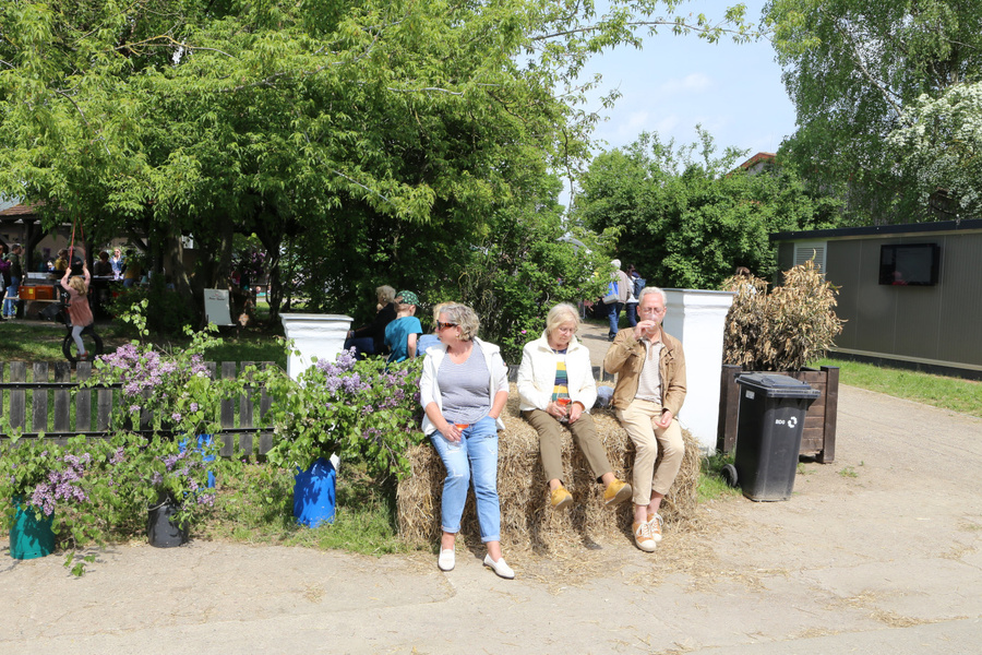 KI generiert: Drei Menschen sitzen auf Strohballen, umgeben von grünen Bäumen und lilafarbenen Blüten, im Freien.