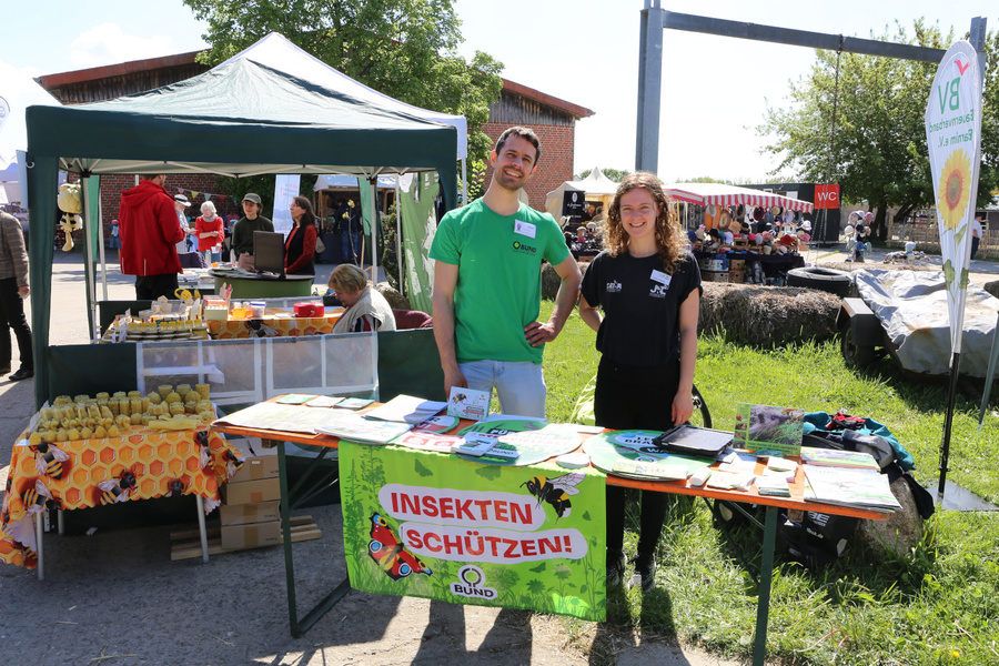 KI generiert: Zwei Personen an einem Infostand mit dem Banner „INSEKTEN SCHÜTZEN!“ von BUND bei einem Outdoor-Event.