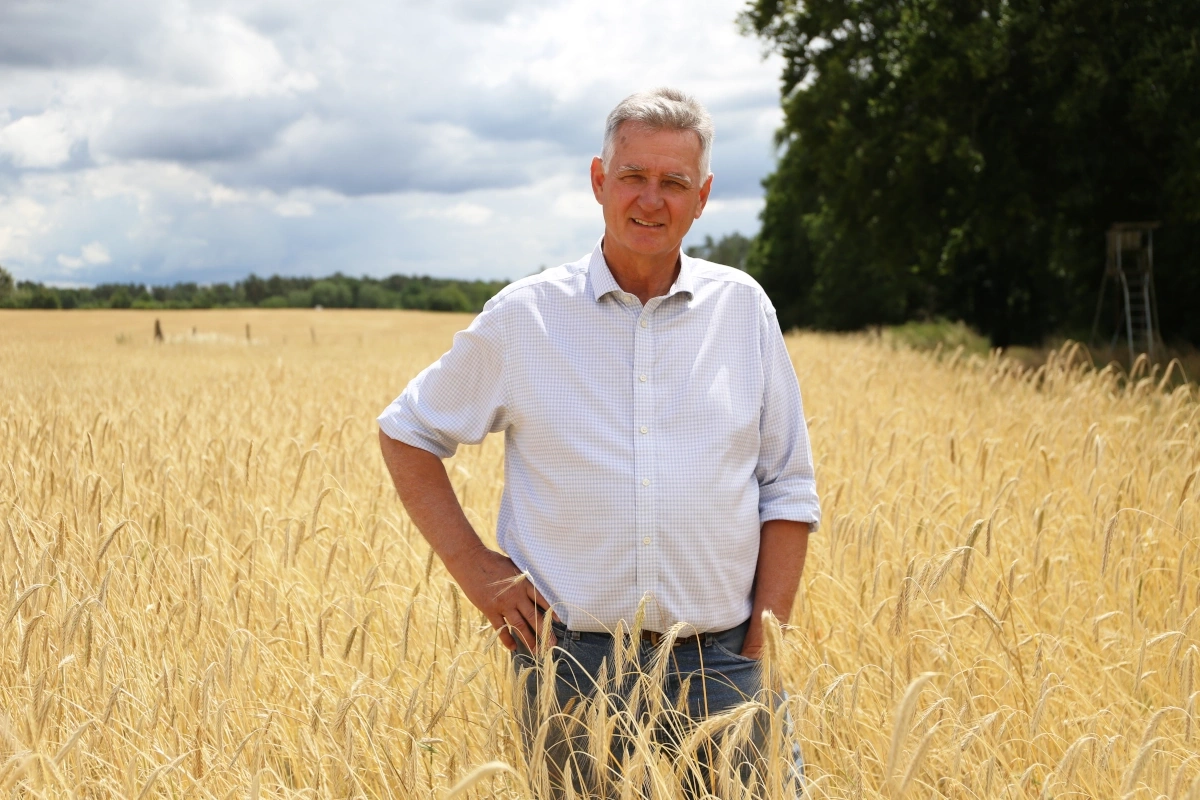 KI generiert: Ein Mann steht in einem Weizenfeld, umgeben von goldenen Ähren und Bäumen im Hintergrund. Kein Text vorhanden.